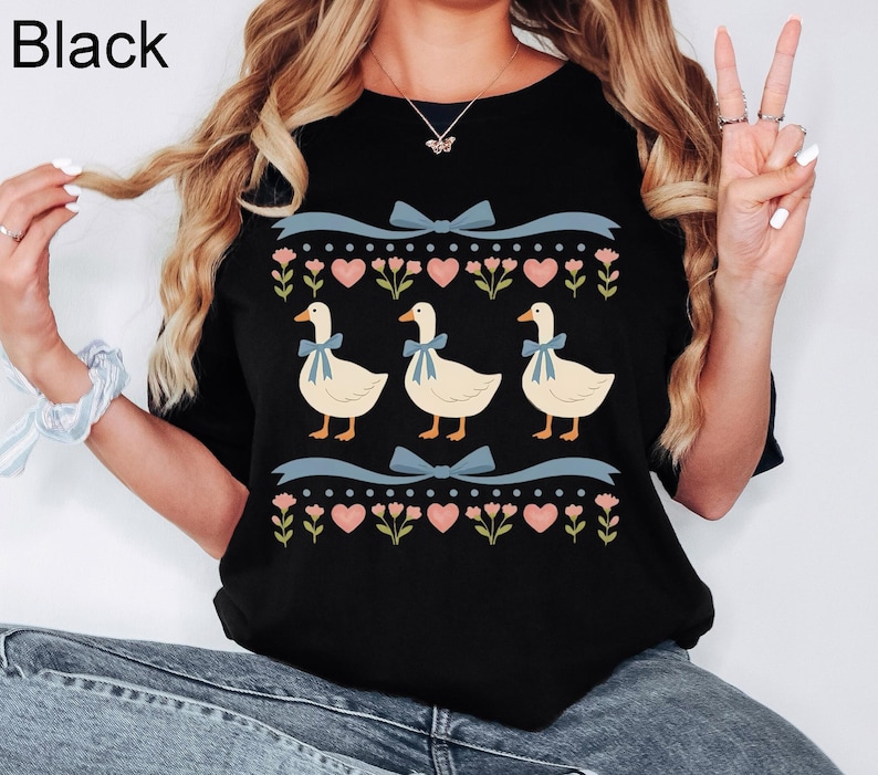 Pode incluir: Camiseta preta com um design l&uacute;dico com tr&ecirc;s gansos usando la&ccedil;os azuis, em um fundo de cora&ccedil;&otilde;es rosa, flores e fitas azuis. A palavra "Black" est&aacute; impressa no canto superior esquerdo.