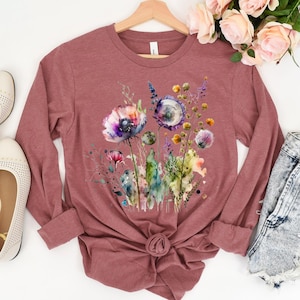 May include: A long-sleeved, mauve-colored shirt featuring a watercolor floral design. The design includes purple, blue, and yellow flowers with green stems and leaves. The shirt is knotted at the waist and hangs on a wooden hanger.