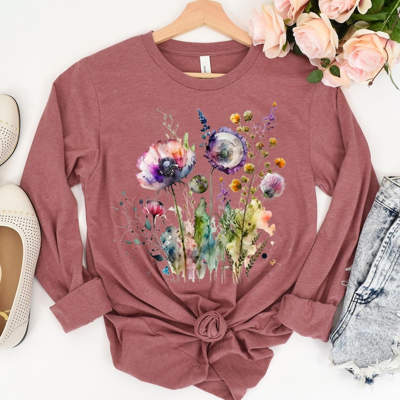 May include: A long-sleeved, mauve-colored shirt featuring a watercolor floral design. The design includes purple, blue, and yellow flowers with green stems and leaves. The shirt is knotted at the waist and hangs on a wooden hanger.