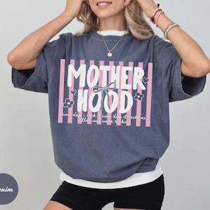 May include: Denim blue short-sleeved t-shirt with the word "MOTHERHOOD" in white block letters, framed by pink vertical stripes. The shirt also features a bow, flowers, and the text "some days it rocks me, other way we're rockin".