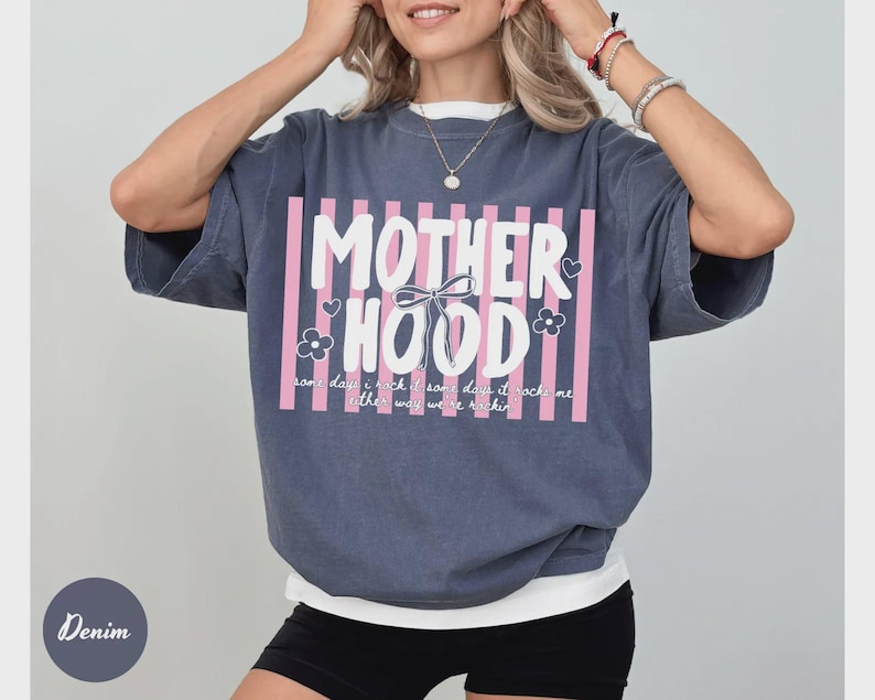 May include: Denim blue short-sleeved t-shirt with the word "MOTHERHOOD" in white block letters, framed by pink vertical stripes. The shirt also features a bow, flowers, and the text "some days it rocks me, other way we're rockin".