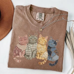 May include: A brown Comfort Colors t-shirt featuring four smiling cat appliqu&eacute;s in various patterns and colors. The cats are arranged in a row, with a paisley, plaid, floral, and patchwork design. A silver necklace with a pendant is draped on the shirt.