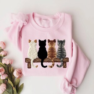 May include: A pale pink sweatshirt with an embroidered design of four cats on a wooden bench. The cats are white, black, brown, and grey. The sweatshirt has long sleeves and a crew neck.