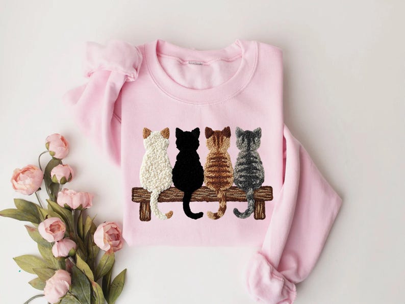 May include: A pale pink sweatshirt with an embroidered design of four cats on a wooden bench. The cats are white, black, brown, and grey. The sweatshirt has long sleeves and a crew neck.