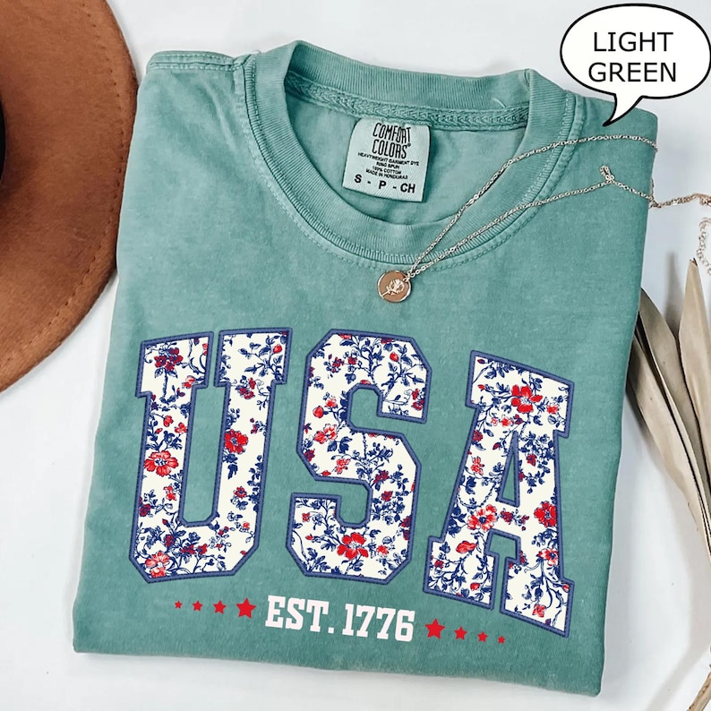 May include: Light green t-shirt with the word "USA" in a floral pattern and the text "EST. 1776". A silver necklace with a pendant is draped across the shirt. The shirt is folded and placed on a surface.
