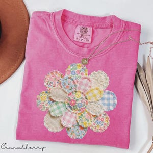 May include: A pink Comfort Colors t-shirt with a floral applique. The applique is made of various patterned fabric circles, including floral, gingham, and solid colors. A gold necklace with a pendant rests on the shirt. The word "Crunchberry" is written in cursive at the bottom.