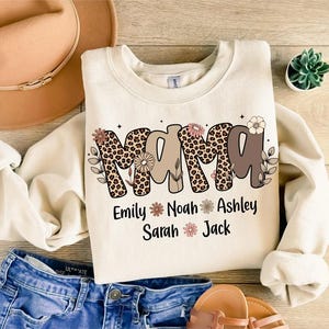 May include: Cream-colored sweatshirt with the word "MAMA" in a mix of leopard print and brown lettering, decorated with floral accents. Below the word are the names Emily, Noah, Ashley, Sarah, and Jack.