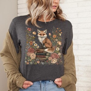 K&ouml;nnte beinhalten: Ein dunkel anthrazitfarbenes T-Shirt mit einem Grafikdruck eines Fuchses, der auf einem Stapel B&uuml;cher sitzt, umgeben von einer Vielzahl bunter Blumen in Orange-, Rosa- und Cremet&ouml;nen. Das Motiv ist mittig auf der Vorderseite platziert.