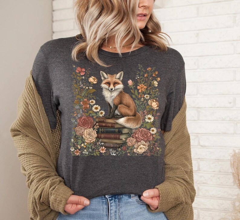 K&ouml;nnte beinhalten: Ein dunkel anthrazitfarbenes T-Shirt mit einem Grafikdruck eines Fuchses, der auf einem Stapel B&uuml;cher sitzt, umgeben von einer Vielzahl bunter Blumen in Orange-, Rosa- und Cremet&ouml;nen. Das Motiv ist mittig auf der Vorderseite platziert.