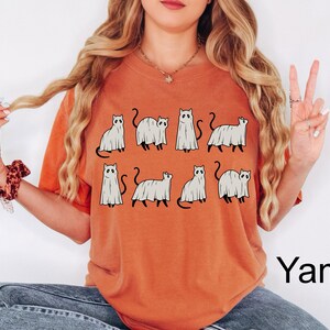 May include: A burnt orange t-shirt with a graphic of multiple white ghost cats. The text "Yam" is in the bottom right corner of the image.