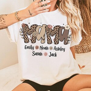 May include: White t-shirt with the word "Mama" in a leopard print design, floral accents, and the names Emily, Noah, Ashley, Sarah, and Jack. The shirt is short-sleeved.