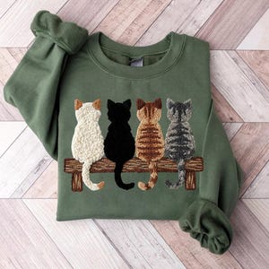 May include: An olive green sweatshirt with an embroidered design of four cats on a wooden bench. The cats are white, black, brown, and grey. The sweatshirt has long sleeves and a crew neck.