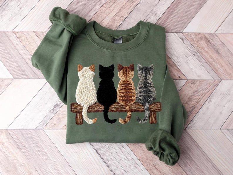 May include: An olive green sweatshirt with an embroidered design of four cats on a wooden bench. The cats are white, black, brown, and grey. The sweatshirt has long sleeves and a crew neck.