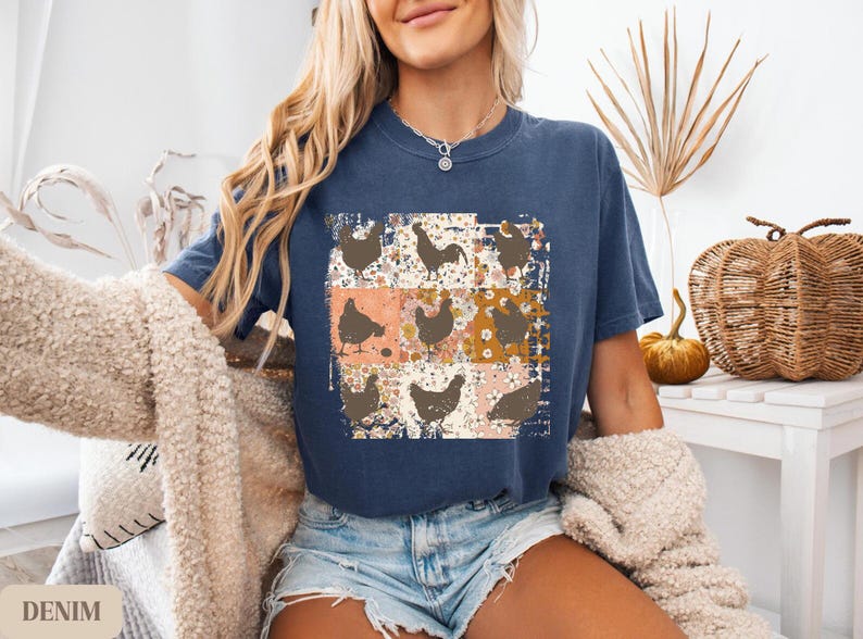 May include: A denim blue t-shirt featuring a patchwork design of chickens and floral patterns. The shirt has a crew neck and short sleeves. The word "DENIM" is visible in the bottom left corner of the image.