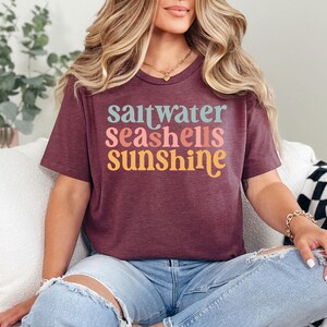 May include: Burgundy t-shirt with the words "saltwater seashells sunshine" in teal, pink, orange, and yellow. The shirt is a crew neck and is worn with blue jeans.