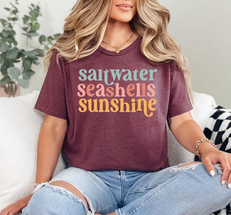 May include: Burgundy t-shirt with the words "saltwater seashells sunshine" in teal, pink, orange, and yellow. The shirt is a crew neck and is worn with blue jeans.
