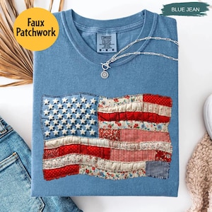 May include: Blue jean colored t-shirt with a faux patchwork American flag design. The flag features white stars and red and white striped fabric. The shirt has the text "Comfort Colors" on the tag and a silver necklace.