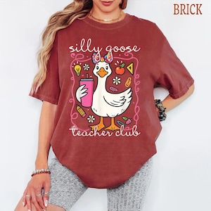 Puede incluir: Camiseta color ladrillo con un dise&ntilde;o de ganso de dibujos animados sosteniendo un vaso rosa, rodeado de ilustraciones escolares. El texto dice "silly goose teacher club". La camiseta es de estilo casual.