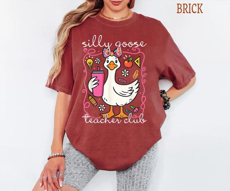 Puede incluir: Camiseta color ladrillo con un dise&ntilde;o de ganso de dibujos animados sosteniendo un vaso rosa, rodeado de ilustraciones escolares. El texto dice "silly goose teacher club". La camiseta es de estilo casual.