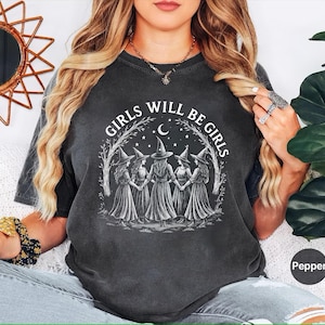 May include: Dark grey t-shirt with the text "GIRLS WILL BE GIRLS" in a circular design. The design features five figures in witch hats holding hands under a crescent moon and stars. The shirt is a Pepper colour.