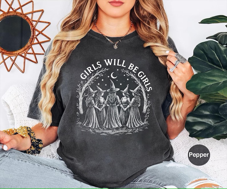 May include: Dark grey t-shirt with the text "GIRLS WILL BE GIRLS" in a circular design. The design features five figures in witch hats holding hands under a crescent moon and stars. The shirt is a Pepper colour.