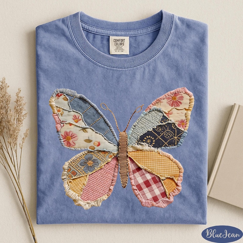 May include: A light blue t-shirt with a large butterfly applique. The butterfly wings are made of various patterned fabrics in shades of pink, blue, yellow, and red. The shirt has a crew neck and a tag that says "Comfort Colors".