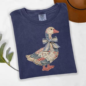 Peut inclure: Un t-shirt bleu marine Comfort Colors avec un motif d'oie en patchwork. L'oie a un bec et des pieds rouges, un n&oelig;ud en vichy bleu et blanc, et un corps fait de divers tissus &agrave; motifs. Un chapeau marron est visible en arri&egrave;re-plan.
