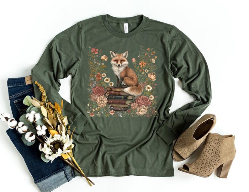 K&ouml;nnte beinhalten: Ein olivgr&uuml;nes Langarmshirt mit einem Grafikdruck eines Fuchses, der auf einem Stapel B&uuml;cher sitzt und von Blumen in Rot-, Orange- und Gelbt&ouml;nen umgeben ist. Das Oberteil verf&uuml;gt &uuml;ber einen Rundhalsausschnitt und lange &Auml;rmel.