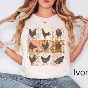 May include: Ivory-colored t-shirt featuring a grid of brown chicken silhouettes on floral and patterned squares. The text "Ivory" is displayed in a black, bold font at the bottom right of the image. The shirt is a casual, everyday wear item.