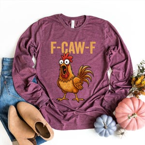 May include: A heathered maroon long-sleeve shirt featuring a cartoon illustration of a surprised-looking orange chicken with wide eyes and an open beak. Above the chicken, the text F-CAW-F is printed in a bold, light-colored sans-serif font.