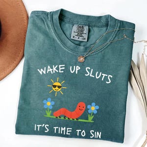 Puede incluir: Camiseta Comfort Colors de color verde azulado con el texto "WAKE UP SLUTS IT'S TIME TO SIN". El gr&aacute;fico incluye un sol sonriente con gafas de sol, un gusano rojo y flores azules. Un collar de oro est&aacute; drapeado sobre la camisa.