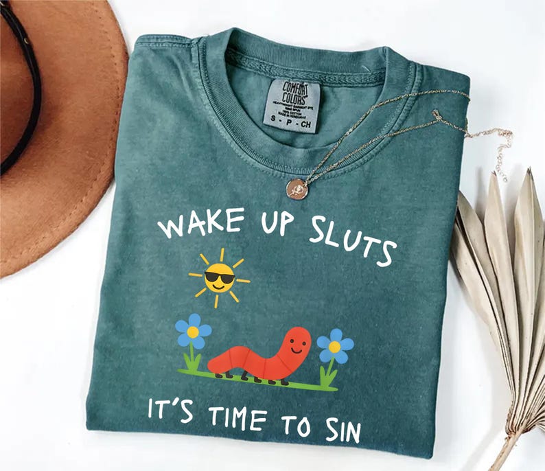 Puede incluir: Camiseta Comfort Colors de color verde azulado con el texto "WAKE UP SLUTS IT'S TIME TO SIN". El gr&aacute;fico incluye un sol sonriente con gafas de sol, un gusano rojo y flores azules. Un collar de oro est&aacute; drapeado sobre la camisa.
