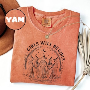 May include: A coral-coloured long-sleeve shirt with the text "GIRLS WILL BE GIRLS" in black, featuring a graphic of five witches. The shirt has a crew neck and a small tag on the upper back. A gold necklace with a pendant is draped on the shirt.