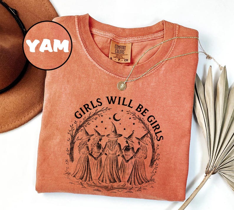 May include: A coral-coloured long-sleeve shirt with the text "GIRLS WILL BE GIRLS" in black, featuring a graphic of five witches. The shirt has a crew neck and a small tag on the upper back. A gold necklace with a pendant is draped on the shirt.