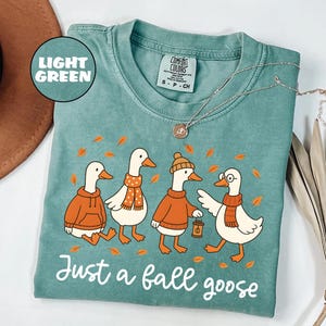 Puede incluir: Camiseta verde claro Comfort Colors con una ilustraci&oacute;n de cuatro gansos de dibujos animados con ropa de oto&ntilde;o. Los gansos llevan su&eacute;teres, bufandas y sombreros, con el texto "Just a fall goose" en escritura blanca. Un collar plateado est&aacute; sobre la camiseta.