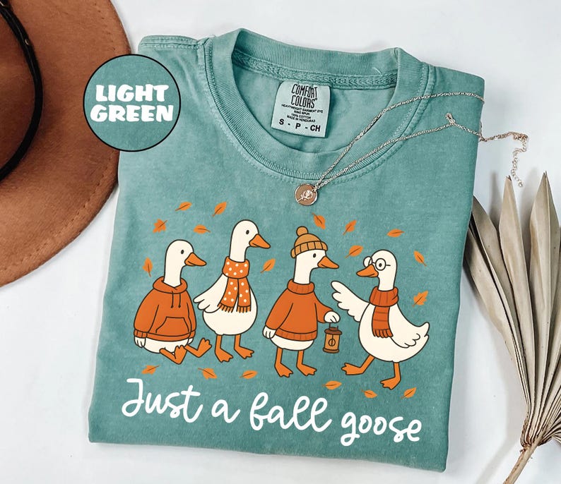 Puede incluir: Camiseta verde claro Comfort Colors con una ilustraci&oacute;n de cuatro gansos de dibujos animados con ropa de oto&ntilde;o. Los gansos llevan su&eacute;teres, bufandas y sombreros, con el texto "Just a fall goose" en escritura blanca. Un collar plateado est&aacute; sobre la camiseta.