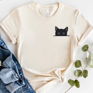 May include: A cream-colored short-sleeve t-shirt featuring a black graphic of a cat head peeking over a horizontal line on the upper left chest. The shirt has a crew neckline and is displayed alongside blue denim fabric and a green leafy branch.