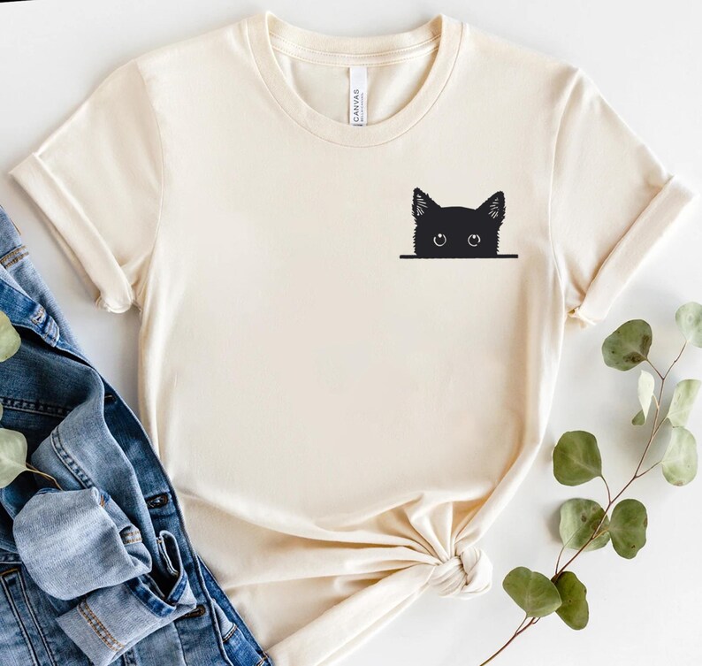 May include: A cream-colored short-sleeve t-shirt featuring a black graphic of a cat head peeking over a horizontal line on the upper left chest. The shirt has a crew neckline and is displayed alongside blue denim fabric and a green leafy branch.