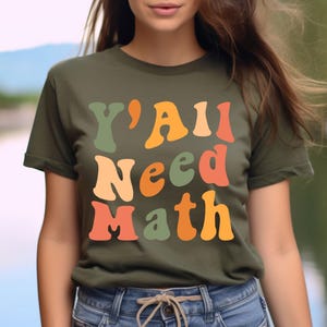 K&ouml;nnte beinhalten: Ein gr&uuml;nes T-Shirt mit dem Text "Y'all Need Math" in bunten Retro-Lettern.