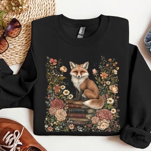 K&ouml;nnte beinhalten: Ein schwarzes Langarm-Sweatshirt mit einem zentralen Motiv eines Fuchses, der auf einem Stapel B&uuml;cher sitzt, umgeben von einem farbenfrohen Arrangement aus verschiedenen Blumen und Bl&auml;ttern in Rosa-, Orange-, Wei&szlig;- und Gr&uuml;nt&ouml;nen.