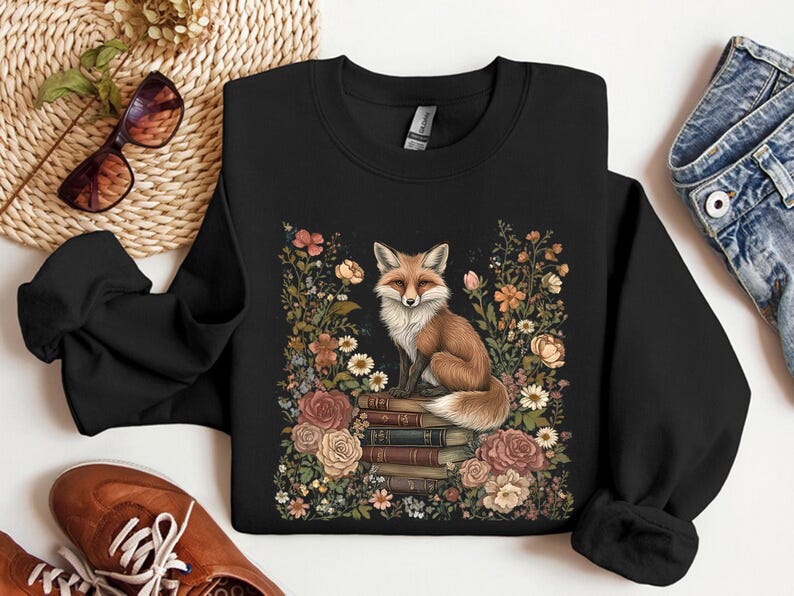 K&ouml;nnte beinhalten: Ein schwarzes Langarm-Sweatshirt mit einem zentralen Motiv eines Fuchses, der auf einem Stapel B&uuml;cher sitzt, umgeben von einem farbenfrohen Arrangement aus verschiedenen Blumen und Bl&auml;ttern in Rosa-, Orange-, Wei&szlig;- und Gr&uuml;nt&ouml;nen.