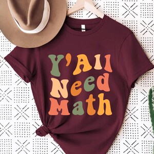 K&ouml;nnte beinhalten: Ein maroonfarbenes T-Shirt mit dem Text "Y'All Need Math" in einer Retro-Schriftart mit bunten Buchstaben.