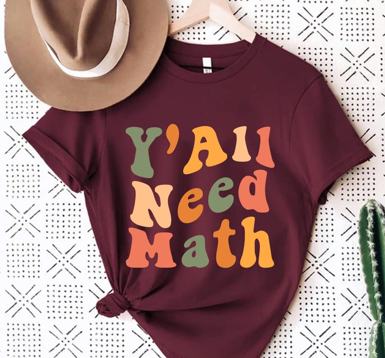 K&ouml;nnte beinhalten: Ein maroonfarbenes T-Shirt mit dem Text "Y'All Need Math" in einer Retro-Schriftart mit bunten Buchstaben.