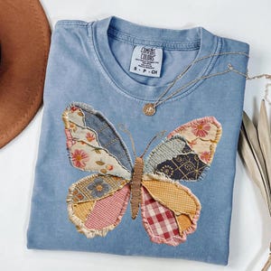 May include: A light blue Comfort Colors t-shirt featuring a large butterfly applique. The butterfly is made of various fabric patches in floral, denim, and plaid patterns. A gold necklace with a pendant rests on the shirt.