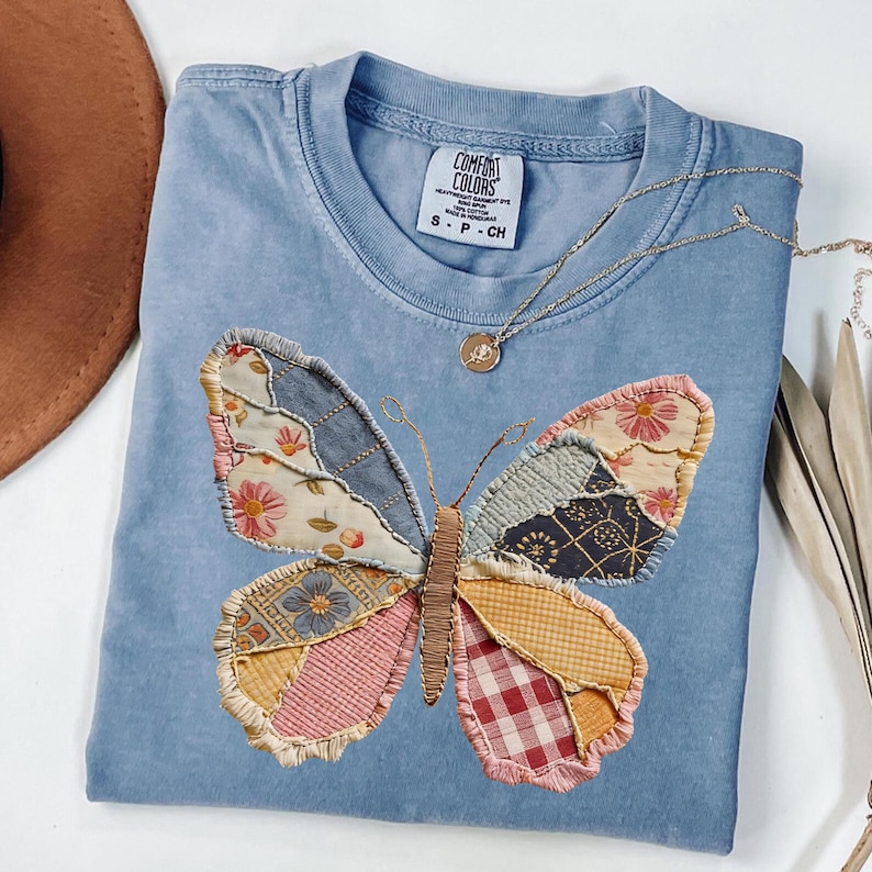 May include: A light blue Comfort Colors t-shirt featuring a large butterfly applique. The butterfly is made of various fabric patches in floral, denim, and plaid patterns. A gold necklace with a pendant rests on the shirt.