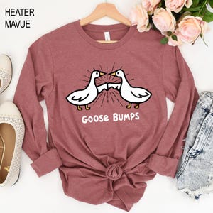 May include: A long-sleeved, mauve-colored shirt featuring a graphic of two white geese with black outlines and yellow feet. The geese are facing each other with a burst design between them, and the words "Goose Bumps" are printed below.