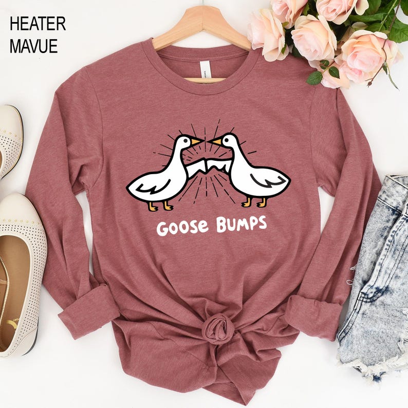 May include: A long-sleeved, mauve-colored shirt featuring a graphic of two white geese with black outlines and yellow feet. The geese are facing each other with a burst design between them, and the words "Goose Bumps" are printed below.