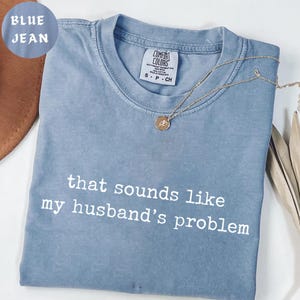 May include: A blue jean Comfort Colors t-shirt with the text "that sounds like my husband's problem" in white. A gold necklace with a pendant rests on the shirt. The shirt is folded and displayed on a neutral surface.