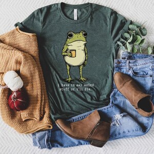 May include: Green t-shirt featuring a cartoon frog holding a mug. The frog is light green and yellow with black outlines. The text "I have to say weird stuff or I'll die" is printed below the frog in a white font. The shirt is paired with blue jeans and brown boots.