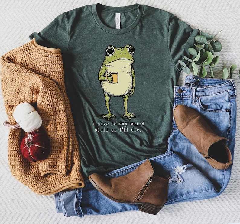 May include: Green t-shirt featuring a cartoon frog holding a mug. The frog is light green and yellow with black outlines. The text "I have to say weird stuff or I'll die" is printed below the frog in a white font. The shirt is paired with blue jeans and brown boots.